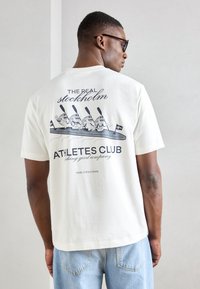 Tricou alb din bumbac cu un imprimeu grafic care prezintă trei personaje de desene animate într-o canoe cu vâsle. Textul include "AFACEREA REALĂ DIN STOCKHOLM" și "CLUBUL ATLETILOR".