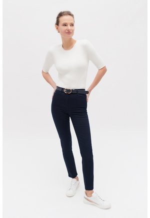 Haut à manches courtes blanc côtelé associé à un pantalon slim-fit bleu marine. Ceinture noire avec boucle dorée, complétée par des baskets blanches avec des accents beiges.