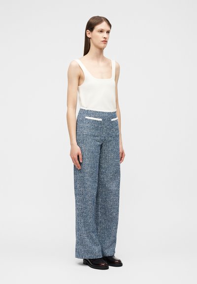 Γυναίκα που φοράει λευκό αμάνικο τοπ, μπλε καρό παντελόνι με φαρδύ πόδι και ψηλή μέση με λευκές τσέπες, και μαύρα παπούτσια, στέκεται ακίνητη.