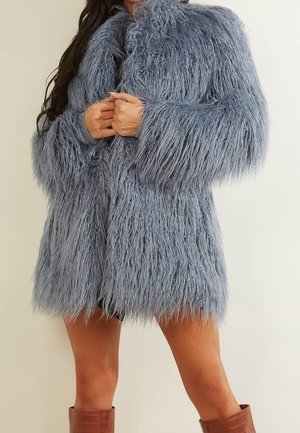 Person trägt einen langen, strukturierten grauen Kunstpelzmantel mit verschränkten Händen, kombiniert mit braunen kniehohen Stiefeln vor einfarbigem Hintergrund.