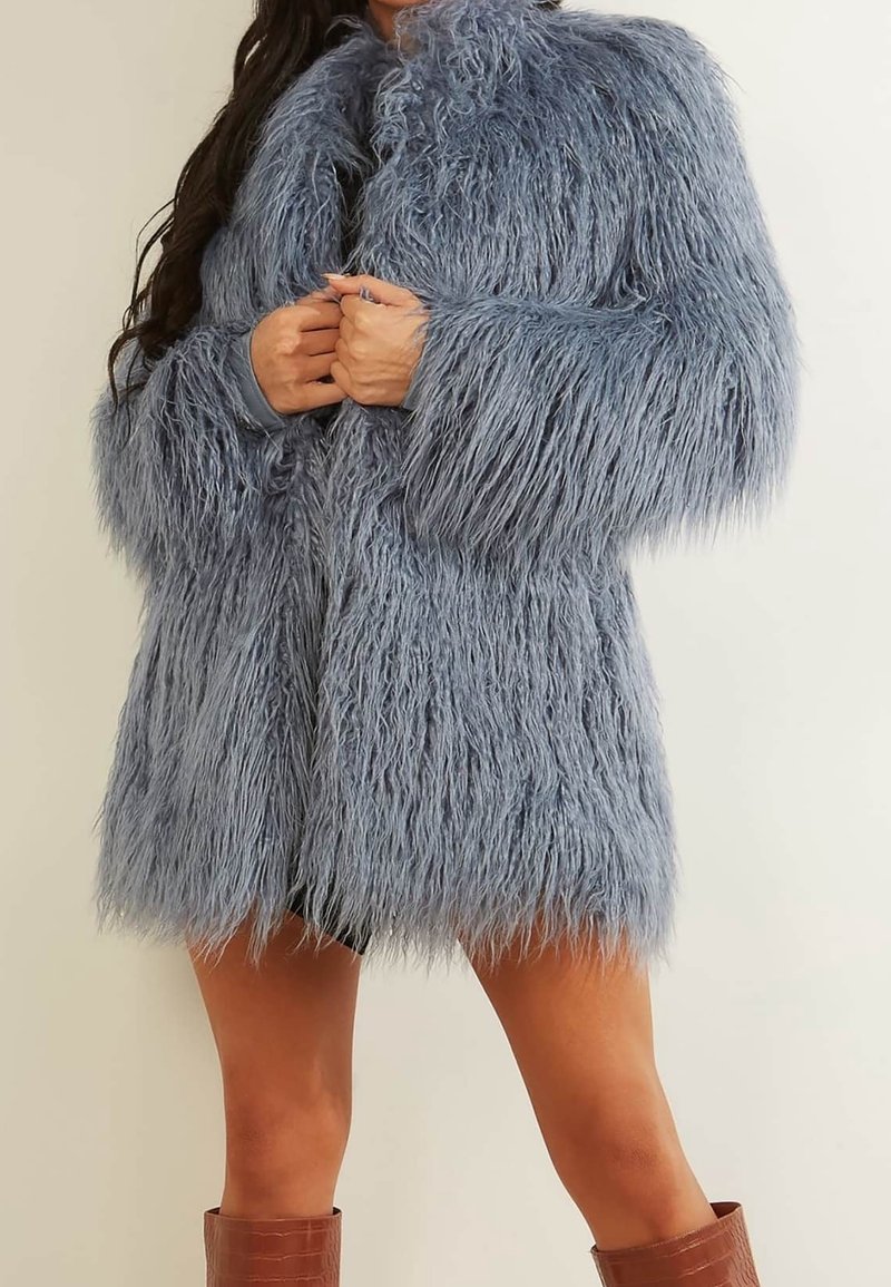 Personne portant un long manteau gris en fausse fourrure texturée avec les mains jointes, associé à des bottes marron montant jusqu'aux genoux sur un fond uni.