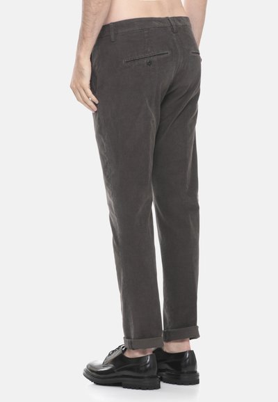 Uomo che indossa pantaloni in velluto a coste grigi scuri e scarpe da cerimonia in pelle nera, visto da dietro su uno sfondo bianco semplice.