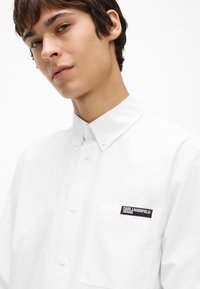 Balta pogājamā kreklā ar klasisku apkakli, ar kreiso krūšu kabatu un melnu zīmola etiķeti. Gluda kokvilnas auduma ar spīdīgu izskatu.