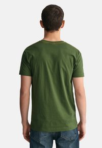 Groene katoenen T-shirt met korte mouwen en een ronde halslijn. Heeft gele stiksels langs de hals. Casual pasvorm, gedragen met blauwe jeans.