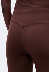 Gros plan d'une personne portant des leggings marron taille haute avec un petit logo sur la ceinture arrière et des détails de coutures visibles.