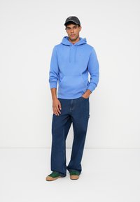 Svetlomodrá mikina s klokaním vreckom, skombinovaná so voľnými tmavomodrými džínsami a zelenými topánkami. Čierna čiapka s logom na prednej strane.