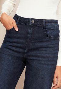 Tumedatest sinisest denimpüksist slim fit stiilis, millel on metallist nööbikinnitus, eespooltaskud ja peened õmbluse detailid.