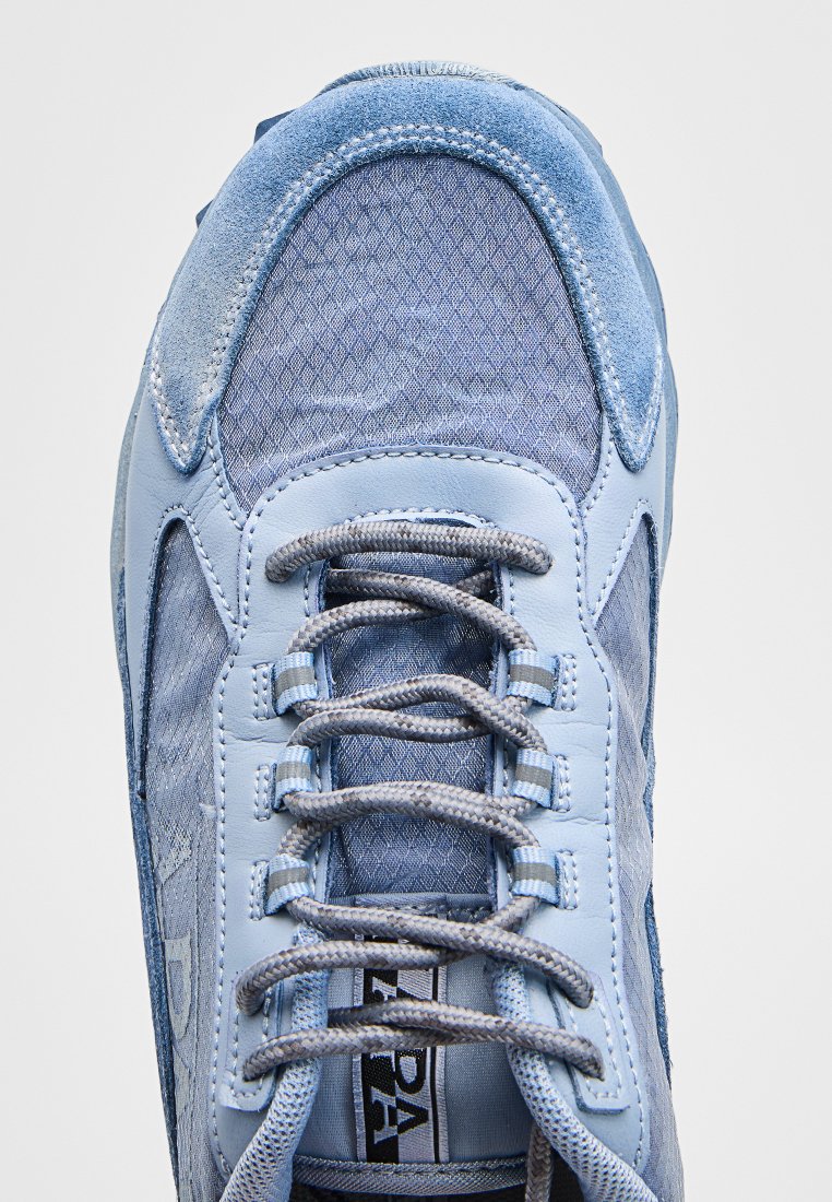 Zapatilla azul claro con paneles de ante y malla, cordones redondos grises atados en patrón cruzado, y una etiqueta de lengua con marca.