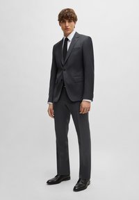Donkergrijs pak met een enkel-breasted blazer en rechte broek. Gecombineerd met een wit overhemd en een zwarte stropdas. Glanzende zwarte dresschoenen.