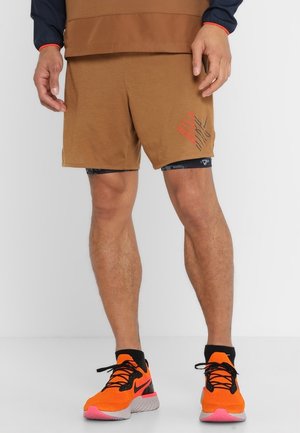 Homme portant un short de sport marron avec un motif superposé noir et orange sur la jambe droite, et des chaussures de course orange vif avec des lacets noirs.