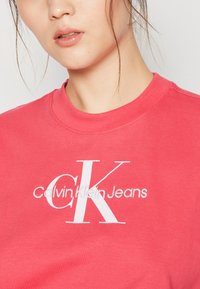 Koralo rožinė džemperis, pagamintas iš minkštos medžiagos, su dideliu baltu "CK" logotipu ir „Calvin Klein Jeans“ tekstu apačioje. Trumpomis rankovėmis ir apvaliu kaklu.