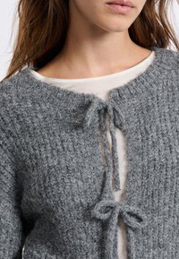Cardigan grigio lavorato a maglia con finitura testurizzata, caratterizzato da un colletto rotondo e chiusure legate sulla parte frontale. Indossato sopra una maglietta leggera.