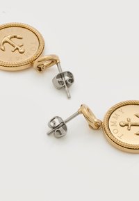 Σκουλαρίκια σε σχήμα νομίσματος με χρυσό τόνο, σχέδιο άγκυρας και κείμενο "TOMMY", με πίσω μέρος καρφί από ασημί μέταλλο σε λευκό φόντο.