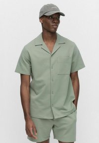 Chemise à manches courtes, de couleur vert clair, avec un motif texturé, fermeture à boutons et poche poitrine, associée à un short assorti et une casquette grise.