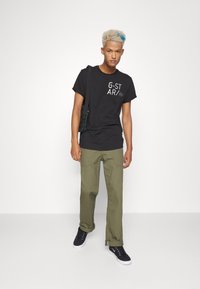 Jovem com cabelo pintado de loiro e mecha azul, vestindo uma t-shirt preta da G-STAR, calças olivas, sapatilhas pretas e a carregar uma pequena mala de ombro.