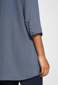 Tunique bleu clair texturée avec manches trois-quarts, munie de poignets à boutons. L'ourlet est légèrement arrondi, offrant une coupe décontractée.