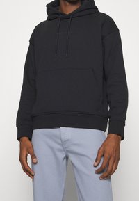 Černá mikina s kapucí, přední kapsou, šňůrkami na stažení a jemným logem. Měkká látka, volný střih, vystavená s světle modrými kalhotami.