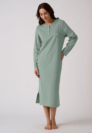 Femme portant une longue chemise de nuit vert pastel avec un col boutonné, une poche à pois avec un nœud et des fentes latérales, debout pieds nus sur un fond uni.