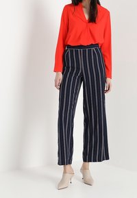 Blouse rouge à manches longues associée à un pantalon large rayé bleu marine et blanc, avec une taille haute et un tissu léger. Chaussures à talons neutres.