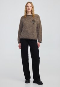 Bruine gebreide trui met een decoratief embleem en kraalaccenten, gecombineerd met zwarte wijde broek en chunky zwarte schoenen.