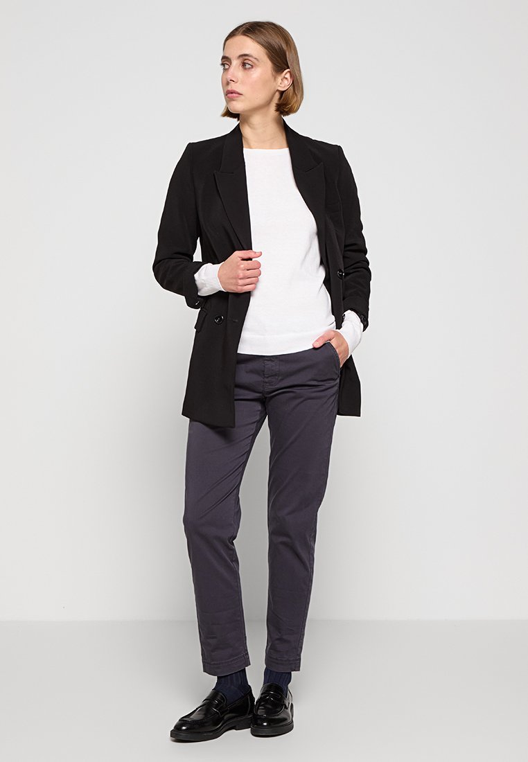 Frau steht, schaut nach links, trägt ein schwarzes Blazer, ein weißes Oberteil, dunkelgraue Hose, schwarze Schuhe und hat kurzes blondes Haar, vor einem schlichten grauen Hintergrund.