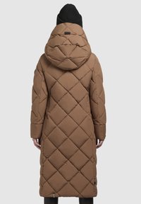 Hnedý dlhý prešívaný puffer kabát s veľkou kapucňou, nosený osobou otočenou chrbtom, skombinovaný s čiernou pletenou čiapkou a čiernymi nohavicami.