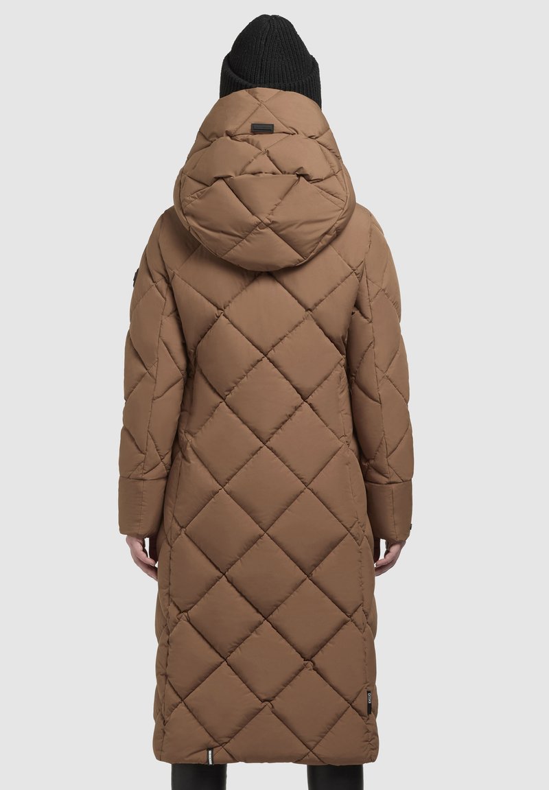 Brauner, langer, gestepptet Puffermantel mit großer Kapuze, getragen von einer Person, die abgewandt steht, kombiniert mit einer schwarzen Strickmütze und schwarzen Hosen.