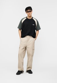 Ung mann iført solbriller, svart og grå kortermet skjorte, beige cargobukser og svarte joggesko, stående med hendene i lommene.