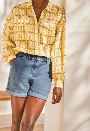 Chemise à carreaux jaunes avec boutons, dotée de deux poches sur la poitrine, associée à un short en denim retroussé. Le tissu semble léger et texturé.