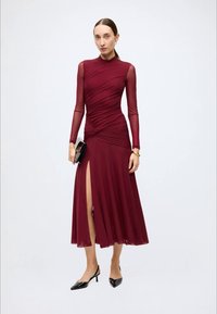 Burgundy long-sleeve dress with a fitted bodice, draped detail, and thigh-high slit. Plain fabric with smooth texture, paired with a black clutch.