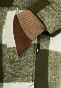 Main à la peau foncée dans la poche d'une veste en laine à carreaux verts et blancs avec un poignet côtelé beige et un bouton-pression sur la manche.
