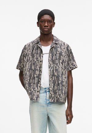 Schlanker dunkelhäutiger Mann, der ein kurzärmeliges schwarz-weiß gestreiftes Hemd mit dem Aufdruck "Karl Lagerfeld Jeans", ein weißes T-Shirt und hellblaue Jeans trägt.