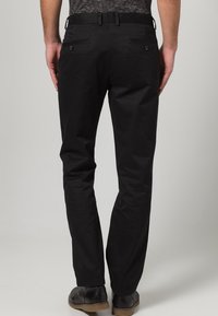 Pantaloni di cotone neri con un design aderente, dotati di tasche posteriori con bottoni e una superficie liscia, indossati con scarpe scure.