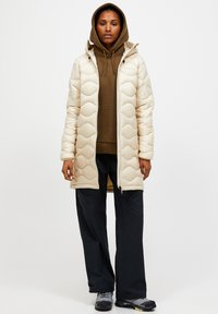 Gevoerde jas in gebroken wit met golvend stikselpatroon, voorzien van een capuchon en een front-rits. Gecombineerd met een bruine hoodie en donkere broek.