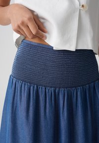 Denim nederdel med en rynket mørkeblå talje, flydende nederste sektion og en glat tekstur. Bæres med en hvid top.