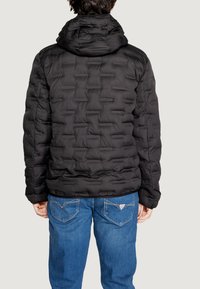 Giacca a vento nera con cappuccio, caratterizzata da una texture trapuntata e polsini elasticizzati. Indossata sopra jeans blu con tasche posteriori visibili.