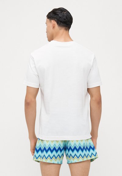 T-shirt blanche à manches courtes avec un col rond ; associée à un short de bain à motif zigzag bleu avec des nuances de vert, bleu et aqua.