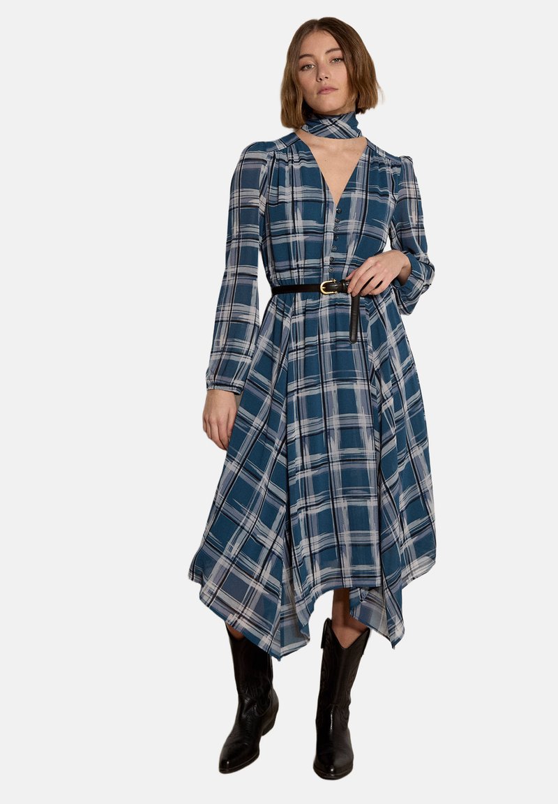 Femme portant une robe à carreaux bleus à manches longues avec un ourlet en mouchoir, une ceinture noire, un foulard assorti au cou et des bottes de cow-boy noires, debout.
