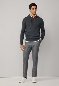 Felpa grigia a maglia con cappuccio, cordino e bottoni, indossata sopra una camicia bianca. Abbinata a pantaloni grigi a righe e sneakers bianche.