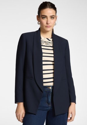 Kvinde med mørkt hår bundet tilbage iført sølv øreringe i form af hoops, marineblå åben blazer, stribet top med blomsterdetalje og blå jeans mod lys baggrund.