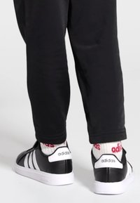 Person trägt schwarze Adidas-Sneaker mit weißen Streifen und weiße Adidas-Socken und steht auf einer hellen Oberfläche.