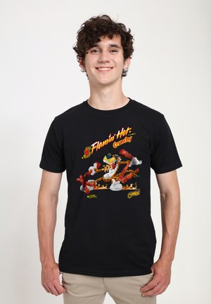 Joven sonriente, vistiendo una camiseta negra con el diseño de "Flamin' Hot Crunchy" Cheetos que incluye una mascota animada y snacks en llamas.