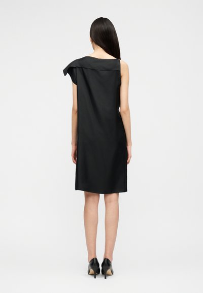 Femme aux longs cheveux noirs portant une robe noire sans manches jusqu'aux genoux avec un col asymétrique, debout de dos, portant des talons noirs.