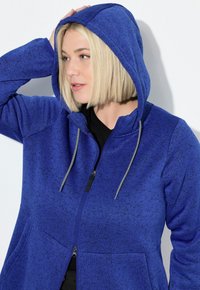 Felpa con zip blu in tessuto testurizzato, cappuccio regolabile, lacci argentati e tasche laterali, caratterizzata da una vestibilità rilassata e accenti scuri.