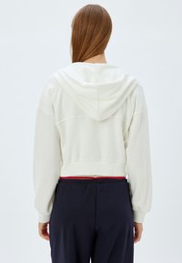 Afskåret, cremet hættetrøje med hætte, ribstrikkede manchetter og kant. Stoffet synes blødt og tekstureret. Båret med mørke bukser med et rødt accent.