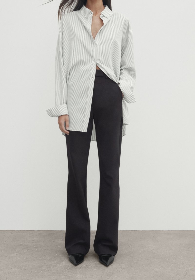Chemise à rayures blanches et grises, assortie à un pantalon large noir et des talons noirs à bout pointu sur un fond neutre.