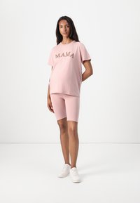 Light pink jersey t-shirt with "MAMA" in a contrasting brown font, paired with matching pink bike shorts. White sneakers complete the outfit.
