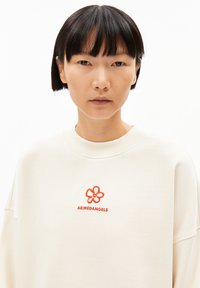 Licht beige oversized sweatshirt van katoen. Voorzien van een rode geborduurde bloem en de merknaam "ARMEDANGELS" eronder. Zachte textuur.