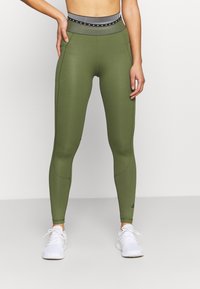 Olivově zelené legíny s vysokým černobílým pruhovaným páskem. Vyrobeny z elastického materiálu, mají hladký povrch a boční kapsy.