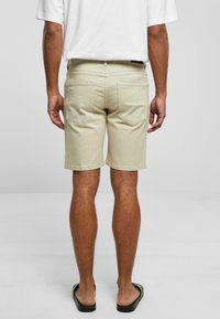 Shorts de algodón beige de corte recto, con dos bolsillos traseros y un acabado con dobladillo. Se llevan con sandalias negras de tipo deslizante.